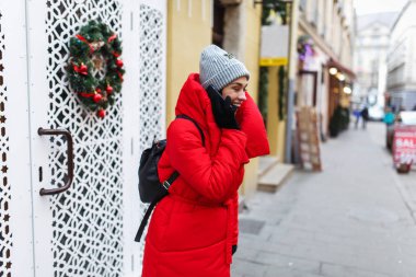 Sokak arka planda gülen kadının portresi cep telefonuyla konuşuyor. Kırmızı kış ceketli kadın kış gününde sokakta duruyor. Noel tatili konsepti. Sağ taraftaki alanı kopyalama