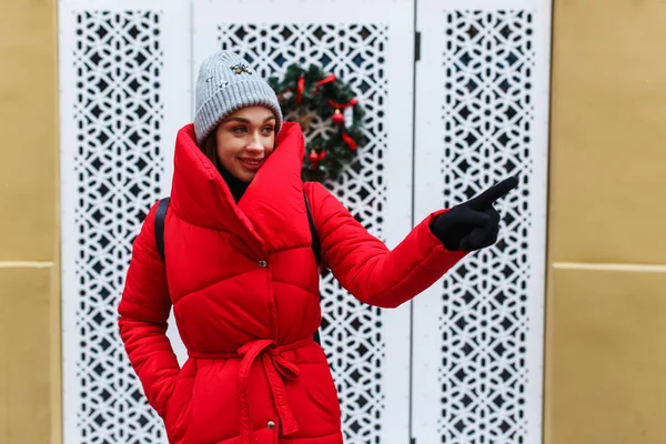 微笑的女人在墙上的背景与圣诞花圈手指指向复制空间 冬天那天 穿着红色冬衣的女人站在大街上 圣诞假期概念 — 图库照片
