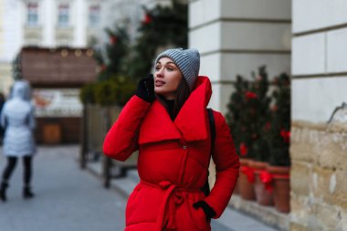 Noel Fuarı cep telefonu konuşurken arka planda ciddi bir kadın portresi. Kırmızı kış ceketli kadın kış gününde sokakta duruyor. Noel tatili konsepti. Sol taraftaki alanı kopyalama