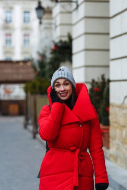 Noel fuarının arka planında ve telefonda konuşan bir kadın portresi. Soğuk kış gününde sokakta yürüyen kırmızı kış ceketli kadın, kış fuarında Noel ağaçları açık havada