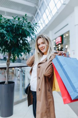 Alışveriş merkezinde kahverengi pelerinli gülümseyen kadın butiklerin arka planında poz lar. Alışveriş merkezinde alışveriş poşetleri olan ve kameraya bakan kadın. Sol taraftaki alanı kopyalama