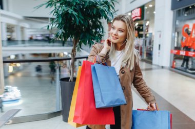 Alışveriş merkezinde kahverengi pelerinli güzel sarışın kadın portresi butiklerin arka planda poz. Alışveriş merkezinde alışveriş poşetleri olan ve kenara bakan bir kadın. Sol taraftaki alanı kopyalama