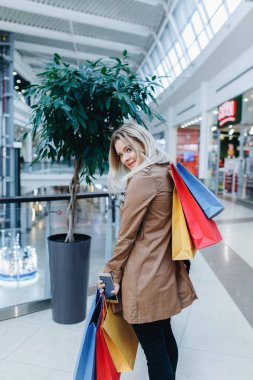 Alışveriş merkezinde kahverengi pelerinli güzel kadın butiklerin arka planında poz. Alışveriş merkezinde alışveriş poşeti olan bir kadın. Kişinin arka tarafı görünümü. Alışveriş, satış, alışveriş merkezi