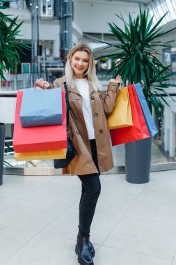 Alışveriş merkezinde kahverengi pelerinli tam uzunlukta sarışın genç kadın butiklerin arka planda poz ve kenara bakıyor. Alışveriş merkezinde alışveriş poşetleri olan güzellik kadını. Alışverişten sonra