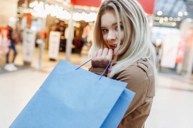 Alışveriş merkezinde kahverengi pelerinli genç kadını yakınbir şekilde butiklerin arka planında poz verirken kameraya bakın. Alışveriş merkezinde alışveriş poşetleri olan güzellik kadını. Sol taraftaki alanı kopyalama