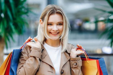 Alışverişten sonra neşeli bir kadın. Bir alışveriş merkezinde kahverengi pelerinli genç kadının portresini kapatın. Alışveriş merkezinde renkli alışveriş poşetleri ile güzellik kadın. Alışveriş, satış, alışveriş merkezi
