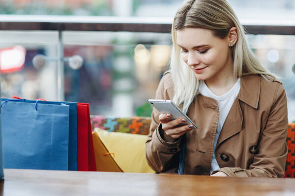 Young woman in brown cloak with shopping bags sitting in cafe at shopping mall. Girl with a phone in her hand. Buying too much concept. Happy shopping. Copy space on the left side