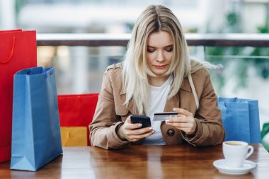 Kız çok para harcadı. Alışveriş merkezindeki kafede alışveriş poşetleri olan sarışın kadın. Bayan bir kredi kartı ve akıllı telefon ile online satın alma. Masada kahve fincanı