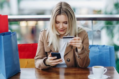 Alışveriş merkezinde kafede oturan alışveriş çantaları ile kahverengi pelerinli güzel sarışın kadın. Bayan bir kredi kartı ve akıllı telefon ile online satın alma. Çok fazla konsept satın alıyorum. Masada kahve fincanı