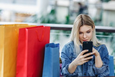 Alışveriş merkezindeki kafede oturan alışveriş poşetleri olan kot ceketli kadın. Elinde cep telefonuyla oynayan kız. Sol taraftaki alanı kopyalayın. Konsept - Alışverişten sonra dinlenmek
