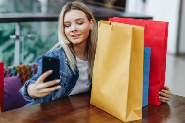 Alışveriş merkezindeki kafede oturan alışveriş poşetleri ile kot ceketli sarışın kadın. Telefonunda sephi yaparken fotoğraflarını çekiyor. Alışverişten sonra rahatlatıcı konsept