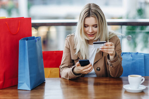 Beautiful blonde woman in brown cloak with shopping bags sitting in cafe at shopping mall. Lady buying online with a credit card and smartphone. Buying too much concept. Coffee cup on table