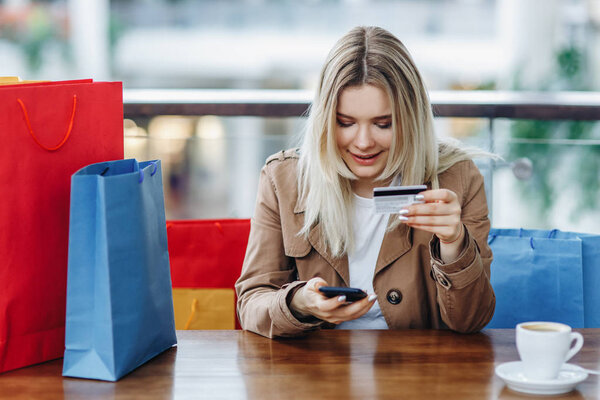 Relaxed blonde woman with shopping bags in cafe at shopping mall. Lady buying online with a credit card and smartphone. Buying too much concept. Coffee cup on table