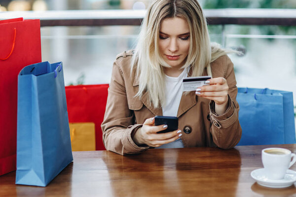 Beautiful woman in brown cloak with shopping bags sitting in cafe at shopping mall. Girl buying online with a credit card and smart phone. Buying too much concept. Coffee cup on table