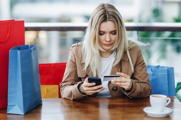 The girl spent a lot of money. Blonde woman with shopping bags in cafe at shopping mall. Lady buying online with a credit card and smartphone. Coffee cup on table