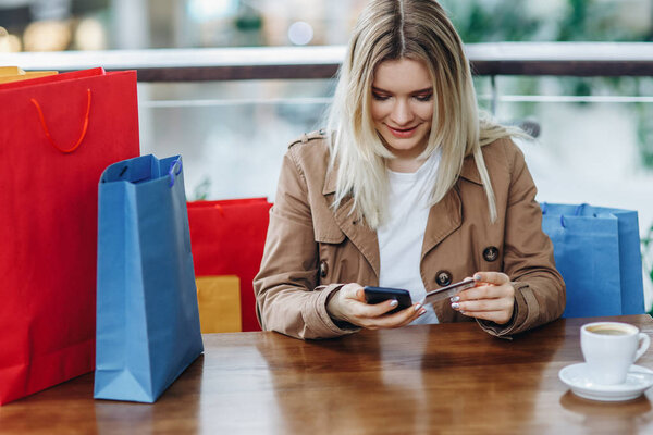 The girl spent a lot of money. Blonde woman in brown cloak with shopping bags in cafe at shopping mall. Lady buying online with a credit card and smartphone. Coffee cup on table
