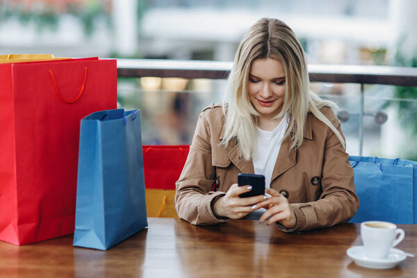 Beautiful blonde woman in brown cloak with shopping bags sitting in cafe at shopping mall. Girl with mobile phone in her hands. Buying too much concept. Coffee cup on table. Time for shopping