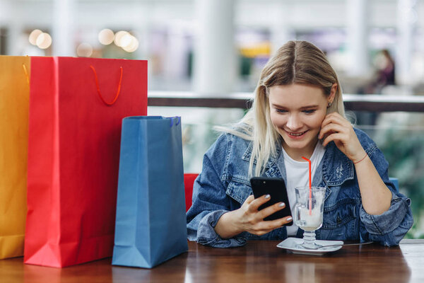 Young woman in a jeans jacket with shopping bags sitting in cafe at shopping mall. Girl with a phone in her hand. Buying too much concept. Happy shopping, copy space