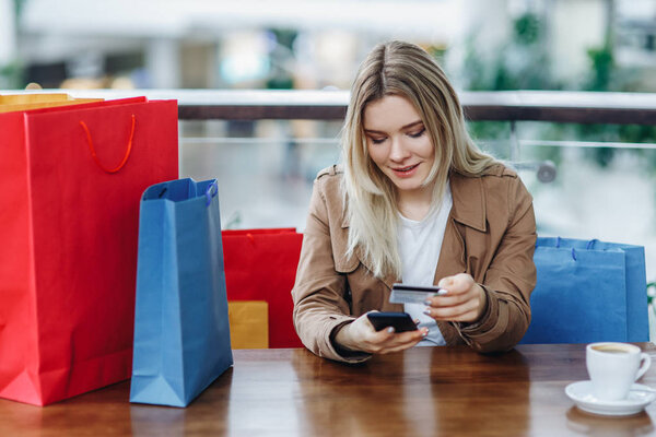 Woman resting after buying and check balance on her bank account. Blonde woman in brown cloak with shopping bags in cafe at shopping mall. Lady buying online with a credit card and smartphone
