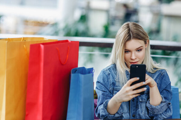 Woman in a jeans jacket with shopping bags sitting in cafe at shopping mall. Girl playing with mobile phone in her hand. Copy space on the left side. Concept - Relaxing after shopping
