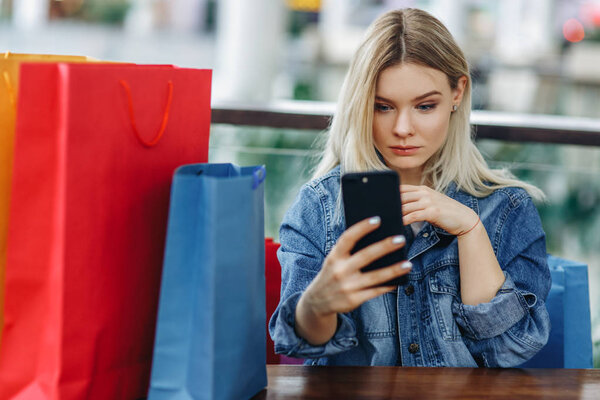 Woman in a jeans jacket with shopping bags sitting in cafe at shopping mall. Girl playing with mobile phone in her hand. Buying too much concept. Concept - Relaxing after shopping
