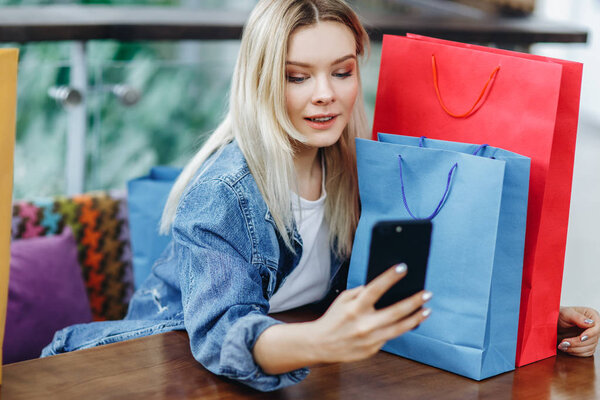 Woman in a jeans jacket with shopping bags sitting in cafe at shopping mall. She is taking pictures of herself on her phone doing a sephi. Concept relaxing after shopping