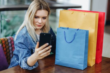 Alışveriş merkezinde kafede oturan alışveriş çantaları ile kot ceket güzel sarışın kadın. Telefonunda sephi yaparken fotoğraflarını çekiyor. Alışverişten sonra rahatlatıcı konsept