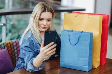 Alışveriş merkezindeki kafede oturan alışveriş poşetleri olan kot ceketli kadın. Telefonunda sephi yaparken fotoğraflarını çekiyor. Masada alışveriş çantaları var. Alışverişten sonra rahatlatıcı konsept