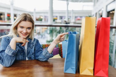 Alışveriş merkezinde kafede oturan alışveriş çantaları ile kot ceket güzel gülümseyen kadın. Kız alışveriş poşetleri üzerinde ve kameraya bakarak gösterir. Mutlu alışveriş, sağ tarafta kopya alanı