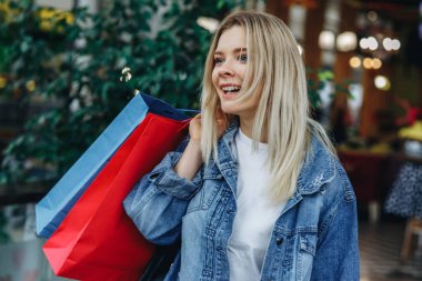 Bulanık alışveriş merkezi arka planda çanta ile bir kot ceket güzel sarışın kız. O alışveriş ten sonra alışveriş çantaları taşıyor ve kenara bakıyor. Satış, mağaza, perakende, tüketici konseptinde model