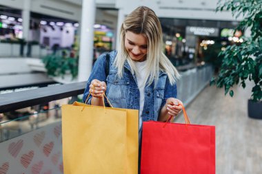 Alışveriş sezonu. Bulanık alışveriş merkezi arka planda renkli alışveriş çantaları ile denim ceket güzel kız. Alışveriş çantasına bakan bir kadın. Sol veya sağ taraftaki alanı kopyalama