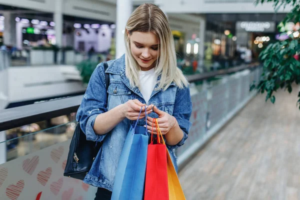 Alışveriş merkezinde alışveriş poşetleri bakarak güzel sarışın genç kadın. Alışveriş merkezinde duran alışveriş çantaları ile kot ceketli güzellik kadını. Sağ taraftaki alanı kopyalama