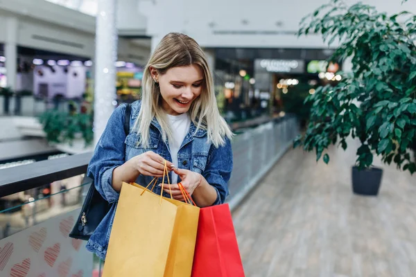 Shopping season. Beautiful laughing girl in a denim jacket with bags on blurred shopping mall background. Urban life and shopogolic concept. Copy space on the right side