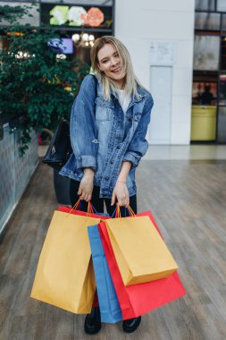 Alışveriş merkezinde ki güzel genç kadın butiklerin arka planında poz lar. Alışveriş merkezinde duran alışveriş çantaları ile kot ceketli güzellik gülümseyen kadın. Alışveriş, satış, alışveriş merkezi