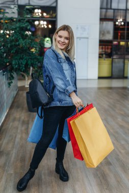 Butiklerin arka planda Moda alışveriş genç sarışın kadın portre. Alışveriş merkezinde duran alışveriş çantaları ile kot ceketli güzellik gülümseyen kadın. Alışveriş, satış, alışveriş merkezi