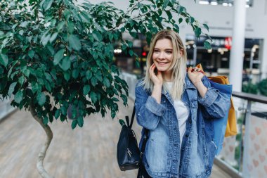 Alışveriş ve ticaret merkezinde zaman harcama zamanı. Elinde renkli alışveriş çantaları ile bir alışveriş merkezinde duran kot ceket güzel gülümseyen kadın. Alışveriş, satış, hediye konsepti