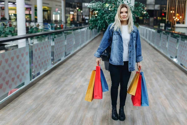 ブティックを背景にショッピングセンターの若い女性の肖像画 ショッピングモールでカラフルなショッピングバッグを持つデニムジャケットの美女 左側のスペースをコピーする — ストック写真
