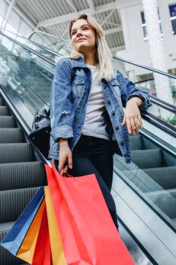 Yürüyen merdiven üzerinde alışveriş merkezinde renkli alışveriş poşetleri ile genç sarışın kadının dikey fotoğraf. Denim ceketli genç alışverişkolik. Satış, mağaza, perakende, tüketici kavramı