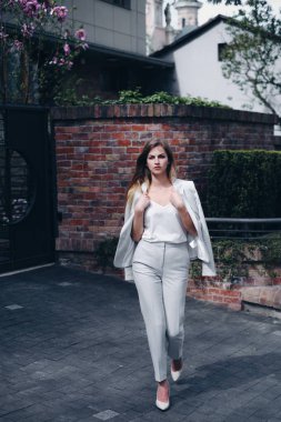 Yüksek ciddi sarışın iş modeli, soluk yumuşak tenli, makyajlı ve yüksek topuklu ayakkabılı büyük muhteşem bahçeli bir ofis yakınında yürüyen. Yeşil bitkileri ve çiçekleri olan rahat modern evler, gri kaldırımlar ve arka planda metalik kapılar. Moda kavramı,