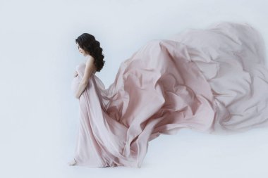 Uzun kıvırcık esmer saçlı, uzun pembe soluk elbiseli, göbeğine dokunan ve aşağıya bakan güzel bir kadın. Hamile, mutlu bir kız, bebek bekliyor ve bekliyor. Annelik ve sevgi kavramı.