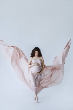 Açık pembe uzun elbiseli, karnını elleriyle tutan güzel hamile bir kadının ön görüntüsü. Güzel hamile bir kadın stüdyoda beyaz bir arka planın önünde duruyor ve aşağıya bakıyor..