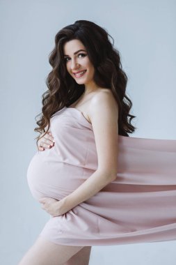 Uzun dalgalı saçlı, çekici hamile bir kadının stüdyo portresi. Uzun kumaşların altında saklanıp sarılıyor, gülümsüyor ve kameraya bakıyor. Sevgi ve ilgi kavramı.