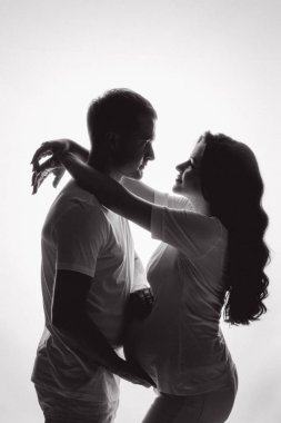 Bebek bekleyen iki ebeveynin stüdyo fotoğrafı. Uzun dalgalı saçlı hamile bir kadın kocasına sarılıyor ve gözlerine bakıyor. Sevgi ve ebeveynlik kavramı.