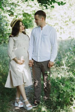 Güzel bir çiftin önünde el ele tutuşup birbirlerine sevgiyle bakıyorlar. Erkek ve hamile kadın, birlikte yeşil parkta yürüyorlar..