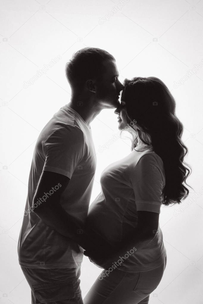 Silhouettes of two marriage couple waiting for baby. Husband touching stomach of his pretty wife and kissing at forehead. Concept of love and parenthood.