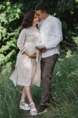 Muhteşem doğada ayakta duran ve rahatlatan güzel bir çiftin ön görüntüsü. Uzun saçlı, açık renk elbiseli, göbeğini tutan hamile bir kadın. Koca güzel öpüyor ve karısının yüzüne dokunuyor.. 