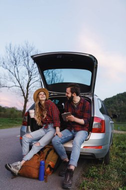 Çift arabanın bagajına oturur ve tabletteki fotoğrafa bakar.