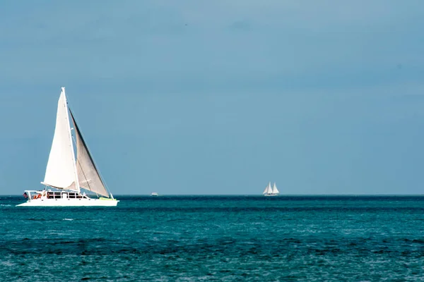 White catamaran sailboat sailing on open blue water with small ketch sailboat in background.