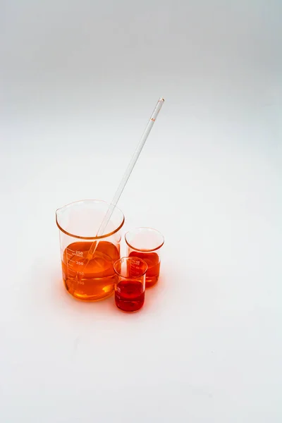 Turuncu ve kırmızı sıvı ile doldurulmuş üç bilim gagalı dikey görüntü ve doğal gölgelerile beyaz bir arka plan üzerinde karıştırma çubuğu