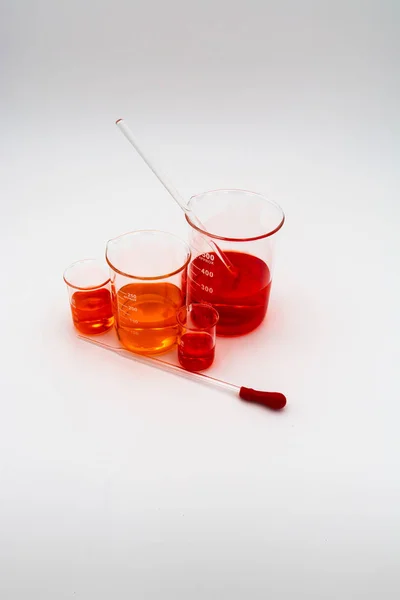 Kırmızı bir ampul ve cam karıştırma çubuğu ile bir damlalık ile beyaz bir arka plan üzerinde turuncu ve kırmızı sıvı ile dolu dört bilim behers dikey görüntü